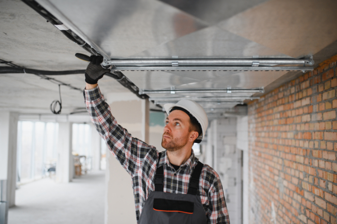 A construction worker in a hard hat and plaid shirt inspects or installs metal pipes on the ceiling of a partially finished building with exposed brick walls, similar to locations where Garage Door Repair Plant City FL services might be needed.