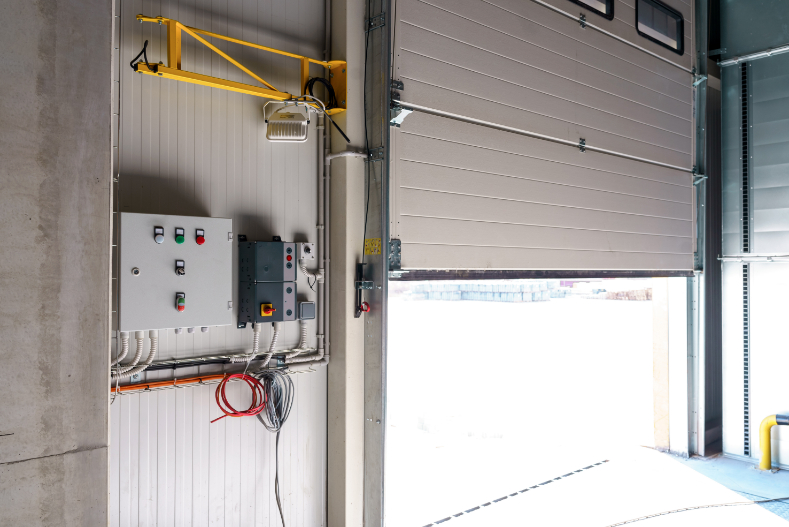 An industrial garage door is partially open, revealing a bright outdoor area. Electrical control panels, buttons, and cables are mounted on the wall inside the building—an everyday scene at a Garage Door Repair Plant City FL facility.
