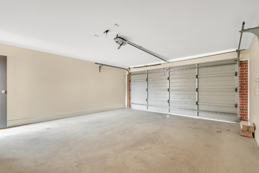 Spacious, empty garage with beige walls, a large closed metal garage door, concrete floor, and a side door to the left. Exposed brick is visible near the garage door—ideal for anyone seeking Garage Door Repair in Plant City FL.